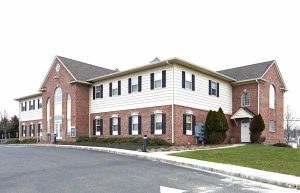 Two-story suburban brick and vinyl office building with white siding, multiple windows, and a parking area in front under an overcast sky.