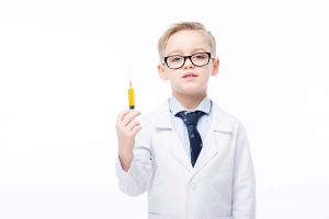 Young boy in glasses and lab coat holding a syringe, posing against a white background.